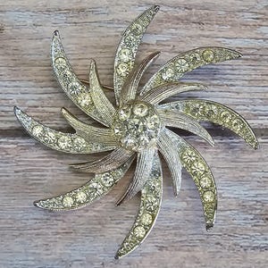 May include: A silver-toned starburst brooch featuring a central cluster of round clear rhinestones surrounded by eight curved, radiating arms. Each arm is embellished with a row of smaller clear rhinestones set into the textured metal surface.