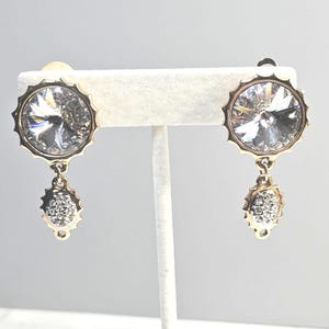 May include: A pair of gold-toned earrings with large, clear, faceted crystal center stones. Each earring features a circular design with a scalloped edge and a smaller, dangling, crystal-studded teardrop shape. The earrings are displayed on a white stand.