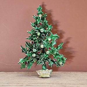 May include: A Christmas tree brooch featuring green enameled holly leaves and a gold-toned base. The tree is decorated with small, round, multi-colored rhinestones in shades of red and iridescent white, arranged throughout the textured foliage.