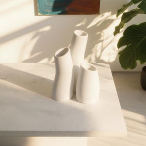 Puede incluir: Tres jarrones de cerámica blanca con formas orgánicas y aberturas anguladas están dispuestos sobre una superficie blanca. Los jarrones tienen un acabado texturizado y varían en altura. La escena está iluminada con luz natural, proyectando sombras.