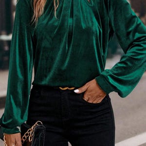 May include: A dark emerald green velvet top with long sleeves and a keyhole detail at the neckline. The top is paired with black pants and a gold belt. A black purse with a gold chain is also visible.