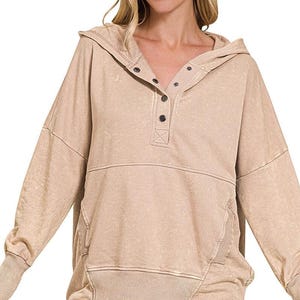 May include: A beige hooded sweatshirt with a button-down neckline. The long-sleeved top has ribbed cuffs and a ribbed hem. The fabric has a slightly faded appearance. The text "ASH MOCHA" is at the bottom right.