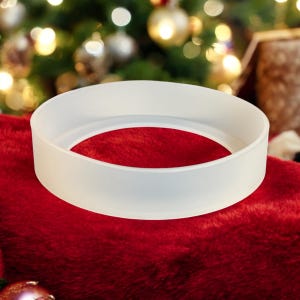 Puede incluir: Un objeto blanco, circular, con un centro hueco, posado sobre una superficie roja y afelpada. El objeto parece estar hecho de un material translúcido. El fondo presenta un árbol de Navidad borroso con adornos.