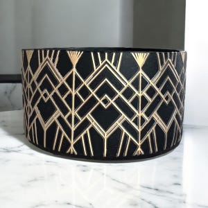 May include: A black cylindrical planter with a gold geometric art deco design. The planter has a matte finish and is suitable for indoor or outdoor use. The design features repeating diamond and linear patterns.