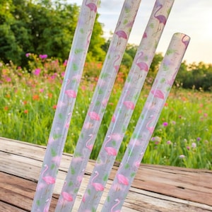 Pode incluir: Quatro palhinhas transparentes com um padrão de flamingo rosa. As palhinhas transparentes apresentam um design repetido de flamingos rosa, folhas verdes e ondas de água. O fundo é uma vista desfocada de um jardim florido.