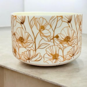 Puede incluir: Un cuenco de cerámica blanco con un diseño floral. El cuenco presenta un patrón repetido de flores y hojas de color dorado. Las flores tienen contornos detallados y están espaciadas uniformemente alrededor del exterior del cuenco. El interior del cuenco es de color blanquecino.