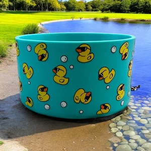 May include: A teal cylindrical container featuring a pattern of yellow cartoon ducks wearing sunglasses and white bubbles. The container is positioned on a sandy shore near a body of water.