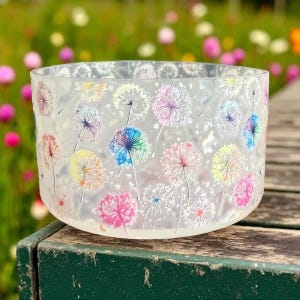 May include: A clear glass bowl with a textured surface, decorated with colorful dandelion designs. The dandelions are in shades of pink, blue, yellow, and purple. The bowl is sitting on a wooden surface.
