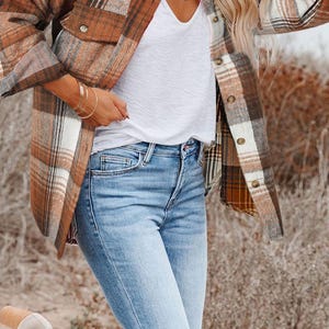 May include: A brown and white plaid flannel shirt jacket with two front pockets, worn over a white V-neck shirt and light blue jeans. The outfit is completed with beige ankle boots. The jacket features a classic button-down design.