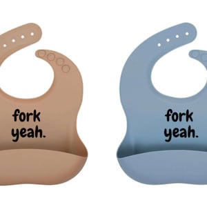 Puede incluir: Dos baberos de silicona para bebé, uno beige y otro azul, con cuello curvo y bolsillo en la parte inferior. Cada babero tiene el texto "fork yeah." en letras negras.