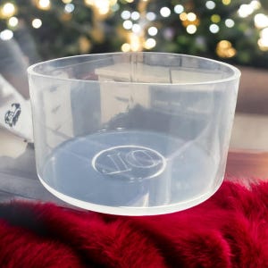 May include: A clear, cylindrical container with a wide opening and a flat, blue-tinted base. The base features a circular design with the letters "JO". The container sits on a red, fluffy surface, with a blurred background of lights.