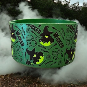 Puede incluir: Un caldero verde con la palabra "WICKED" y gráficos de brujas. El caldero tiene la frase "Defying Gravity" y otro texto. El caldero está rodeado de humo, creando una atmósfera mística.
