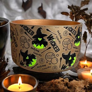 Puede incluir: Un recipiente decorativo, redondo, de color marrón claro con un interior dorado. El exterior presenta la palabra "WICKED" y gráficos de brujas con el texto "Defying Gravity". El recipiente está iluminado con un brillo verde, perfecto para la decoración de Halloween.