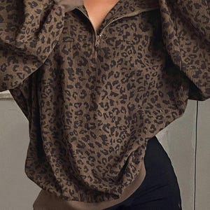 Puede incluir: Jersey de pana marrón con estampado de leopardo. La parte superior presenta cierre de media cremallera, mangas largas y un ajuste holgado. Las mangas y el cuello son de color marrón liso. Combinado con pantalones cortos negros.