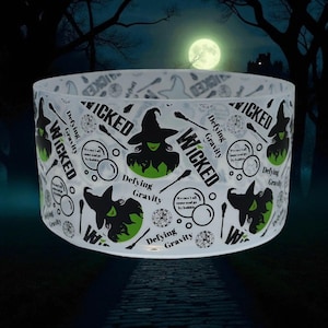 Puede incluir: Un recipiente cilíndrico transparente con temática de Halloween. El diseño presenta siluetas de brujas negras, detalles en verde y la palabra "WICKED" en negrita. La frase "Defying Gravity" también está presente, junto con otros textos y gráficos relacionados con brujas.