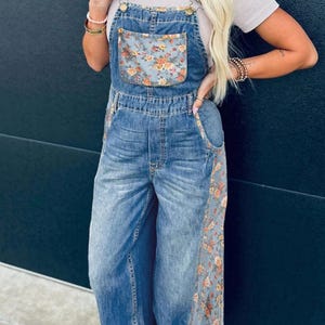 Könnte beinhalten: Blaue Jeans-Latzhose mit einer floral bedruckten Tasche und Seitenteilen. Die Latzhose hat verstellbare Träger und ein weites Bein-Design. Das Blumenmuster umfasst orange, gelbe und rosa Blüten auf blauem Grund. Getragen mit einem hellen Oberteil.