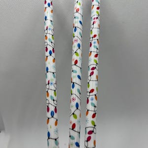 May include: Three white, cylindrical pens with a repeating pattern of colorful Christmas lights and snowflakes. The lights are red, blue, green, and yellow. The pens are standing upright against a white background.