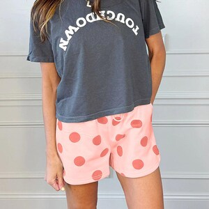 Könnte beinhalten: Dunkelgraues T-Shirt mit dem weißen Wort "TOUCHDOWN", kombiniert mit rosa Shorts mit großen korallenfarbenen Punkten. Das Outfit wird durch ein rosa Visier ergänzt.