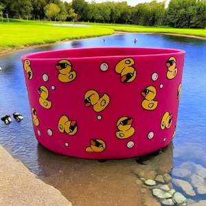 May include: A vibrant pink, cylindrical container adorned with a repeating pattern of yellow rubber ducks sporting sunglasses. The container is positioned on a sandy shore, adjacent to a body of water, with a green grassy area in the background.