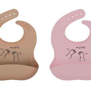 Puede incluir: Dos baberos de silicona para bebé, uno beige y otro rosa, con cuello curvo y bolsillo en la parte inferior. Cada babero tiene la palabra "more" y una ilustración de dos manos. Diseñados para una fácil limpieza.