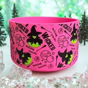 Puede incluir: Un recipiente cilíndrico rosa brillante con un patrón repetido de siluetas de brujas, la palabra "WICKED" y la frase "Defying Gravity". La bruja lleva un sombrero negro y tiene la piel verde. El recipiente es probablemente una taza o un soporte.
