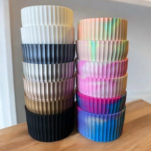 May include: A collection of stacked, cylindrical planters in various colors. The planters have a ribbed design and range in color from white, black, and gold to pastel and vibrant hues, including pink, blue, and a rainbow gradient. They are arranged on a wooden surface.