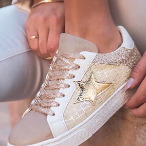 May include: Cream-colored sneakers with a gold star applique. The shoes feature a beige suede toe cap, gold glitter accents on the heel, and gold-toned laces. The side panel has a textured pattern.