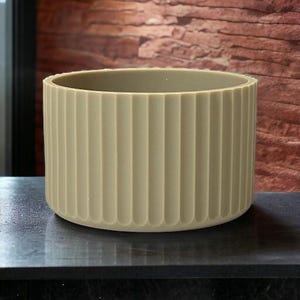 May include: A cylindrical, light beige planter with vertical, ridged detailing. The interior is a slightly darker shade of beige. The planter sits on a dark surface, with a brick-like wall in the background. This decorative planter is suitable for indoor or outdoor use.