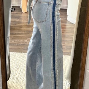 May include: Light wash denim jeans with a wide leg and a dark blue vertical stripe down the side. The stripe has a frayed edge. The jeans are reflected in a mirror.
