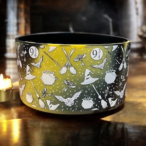 Puede incluir: Contenedor cilíndrico con interior negro y exterior degradado de amarillo a negro. El contenedor está decorado con ilustraciones blancas de un sombrero de bruja, escobas, llaves, búhos y el número 9 3/4.