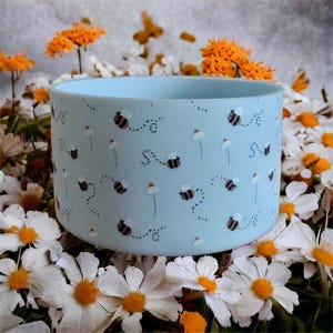 Puede incluir: Un recipiente cilíndrico azul claro decorado con abejas de dibujos animados y trayectorias de vuelo punteadas. Flores blancas y naranjas rodean el recipiente. Las abejas tienen rayas negras y amarillas.