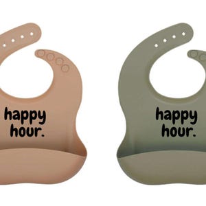Puede incluir: Dos baberos de silicona para bebés, uno en color melocotón apagado y otro en verde apagado. Cada babero tiene un cuello curvo, broches ajustables y un bolsillo para recoger alimentos. Las palabras "happy hour." están impresas en negro.
