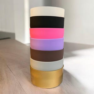 Puede incluir: Una pila de bandas de silicona de colores, incluyendo blanco, negro, rosa, morado, marrón, blanco roto y dorado. Las bandas están apiladas una encima de otra, creando una exhibición visualmente atractiva. Las bandas son probablemente para vasos.