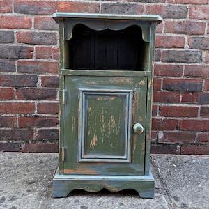 May include: A small, distressed green wooden cabinet with a single door and open shelf. The cabinet features a scalloped top and base, and a round knob. The paintwork has a worn, antique appearance, with blue and orange tones visible.