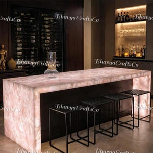 May include: A contemporary bar area with a large, illuminated pink countertop with a marble-like effect. Black metal stools are in front of the counter. A wine rack and shelves with glassware and bottles are in the background.