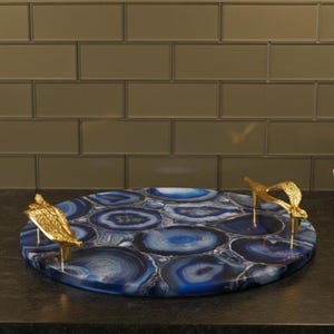 Puede incluir: Una bandeja redonda para servir con un diseño de ágata azul y asas doradas. La bandeja tiene aproximadamente 35 cm de diámetro, con una superficie pulida y asas decorativas. Las asas tienen forma de hojas estilizadas.