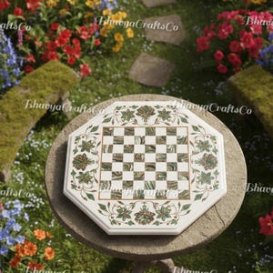 Puede incluir: Tablero de ajedrez octogonal blanco con un diseño floral verde y dorado. El tablero de ajedrez tiene una superficie de juego a cuadros y está colocado sobre una mesa redonda de piedra. El fondo presenta un jardín con flores coloridas.