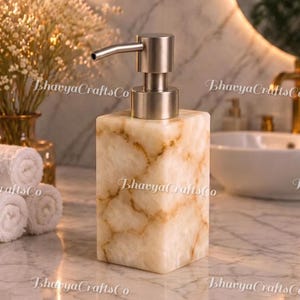 Puede incluir: Un dispensador de jabón rectangular de piedra color crema con vetas marrones y una bomba de acero inoxidable. El dispensador está sobre una superficie de mármol.