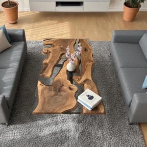 Pode incluir: Uma mesa de centro de madeira com bordas naturais e formato orgânico. A mesa apresenta um preenchimento de resina transparente e está sobre um tapete cinza. Um pequeno vaso com flores e um livro estão sobre a mesa.