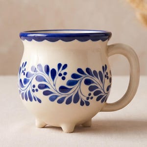May include: Cream-colored ceramic mug with a blue floral design and a scalloped rim. The mug has a rounded body, a curved handle, and three small feet. A woven basket with dried plants is in the background.