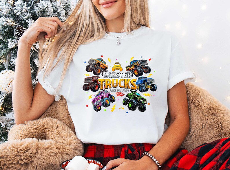 A woman wearing a white t-shirt with a graphic of monster trucks and the text "MONSTER TRUCKS" is sitting in front of a Christmas tree.