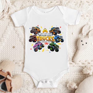 A white baby onesie with a graphic of monster trucks printed on it, placed on a white blanket. There are also a pair of baby shoes and a stuffed animal nearby.