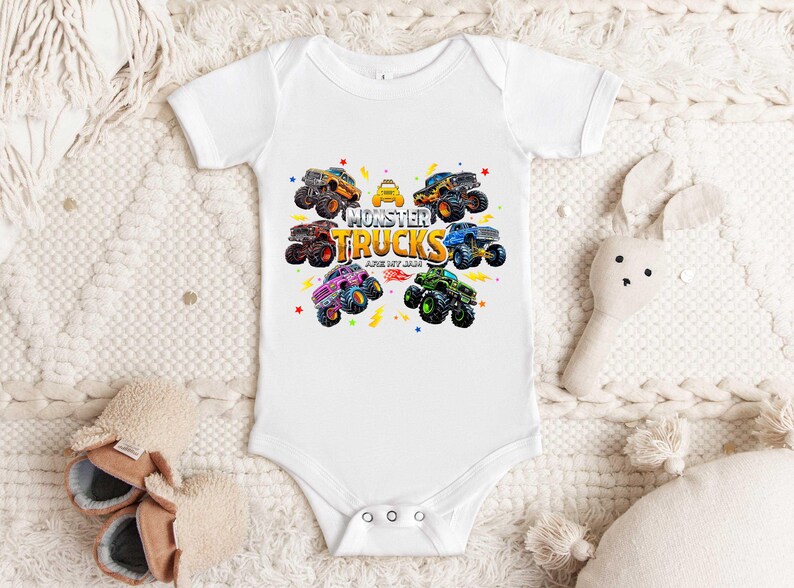 A white baby onesie with a graphic of monster trucks printed on it, placed on a white blanket. There are also a pair of baby shoes and a stuffed animal nearby.