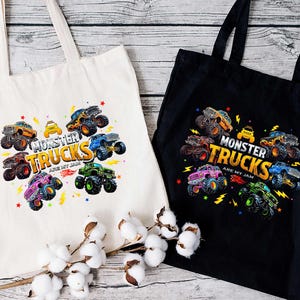 two tote bags with a vibrant design of monster trucks printed on them. The bags are placed on a wooden surface, with a branch of cotton bolls in the foreground.