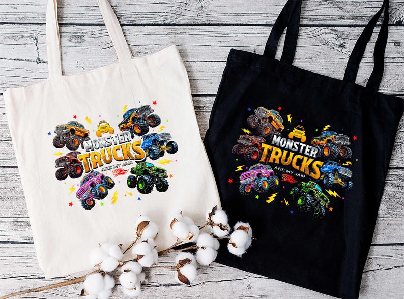 two tote bags with a vibrant design of monster trucks printed on them. The bags are placed on a wooden surface, with a branch of cotton bolls in the foreground.