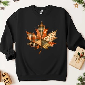 A black sweatshirt with a colorful autumn leaf design on it, surrounded by holiday decorations such as pine branches, pinecones, and a small gift box.