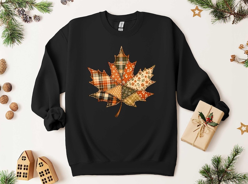 A black sweatshirt with a colorful autumn leaf design on it, surrounded by holiday decorations such as pine branches, pinecones, and a small gift box.