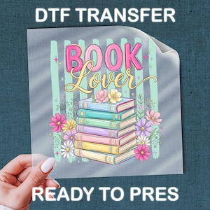 May include: A DTF transfer sheet featuring a stack of colorful books with the text BOOK Lover in pink and gold script. The design includes pastel mint vertical stripes and various pink, yellow, and purple flowers surrounding the book stack.