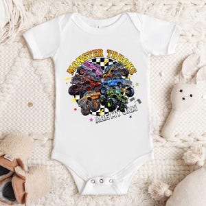 A white baby onesie with a graphic of monster trucks printed on it, along with a stuffed rabbit and a pair of baby shoes.