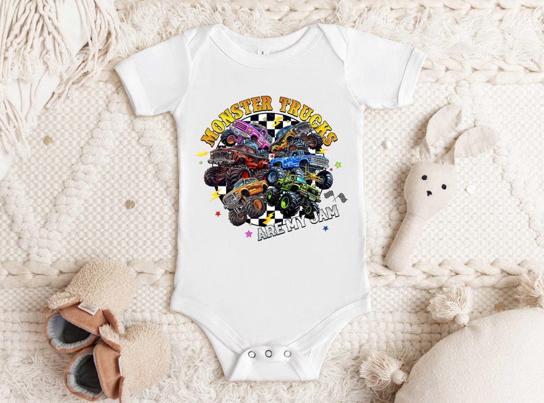 A white baby onesie with a graphic of monster trucks printed on it, along with a stuffed rabbit and a pair of baby shoes.